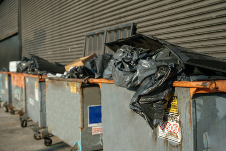 A close-up view of a rusty industrial dumpster overflowing with black trash bags and cardboard. Warning stickers and weathered surfaces highlight the gritty urban setting.の写真素材
