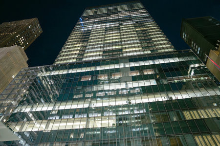 A modern glass skyscraper glows brightly at night in the heart of New York City. The reflection of...