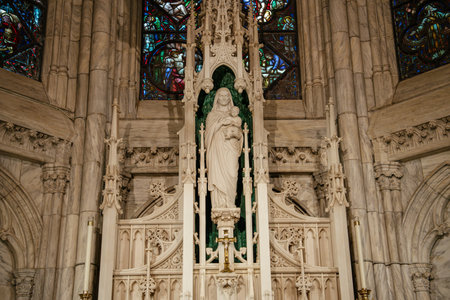 This image features a detailed white marble statue of the Virgin Mary holding the Christ Child, set within a richly carved altar niche at St. Patricks Cathedral in New York City. Stained glass windows and decorative Gothic elements surround the sacred figure.の写真素材