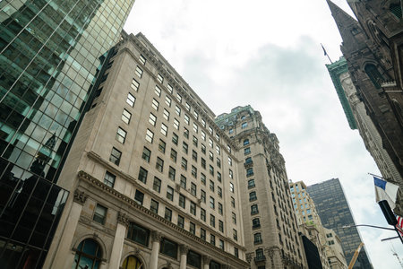 A mix of stone facades and glass buildings rises into a cloudy New York sky. Reflections and...