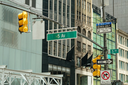 Green Fifth Avenue street signs, yellow traffic lights, and bus lane signage mark a busy New York City intersection. Iconic urban architecture frames the scene in the background.の写真素材