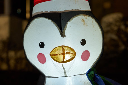 A smiling inflatable penguin with rosy cheeks and a yellow beak glows under night lights. The cute holiday character wears a red Santa hat in a festive scene.の写真素材