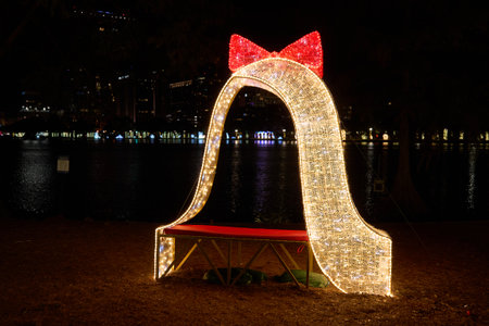 Illuminated arch with a red bow and bench glows beside the city lake at night. Festive holiday decoration with bright LED lights and golden sparkle.の写真素材