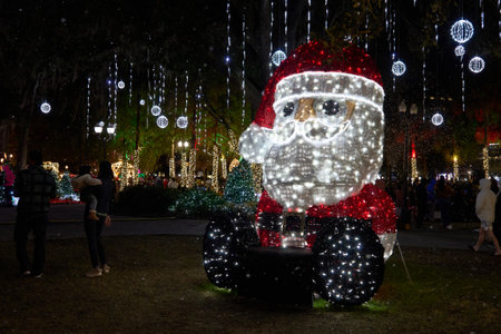 Giant Santa Claus made of LED lights glows in a festive park at night. Christmas decoration with snow, ornaments, and bright sparkle around families.の写真素材