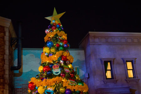 Orlando, Florida, USA - 11.15.2024: A tall Christmas tree is covered with bright yellow garlands, shiny ornaments, and playful cartoon faces. The glowing tree stands between city buildings under the dark night sky.のeditorial素材