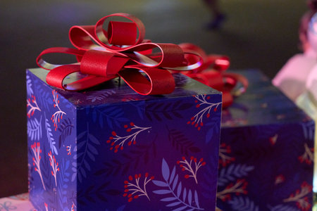 A blue gift box with a large red ribbon bow sits under holiday lights. The wrapped present shows festive colors and glossy paper in a closeup night scene.の写真素材