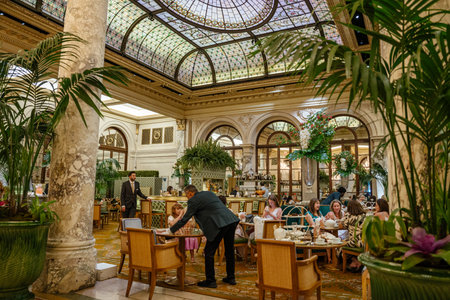 New York City, New York, USA - 08.01.2024: Guests enjoy afternoon tea in a grand hotel lounge with a stained glass dome ceiling and marble columns. The refined interior, floral arrangements, and lush greenery create a sophisticated atmosphere.のeditorial素材