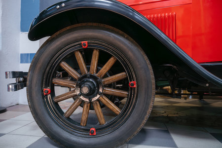 Detailed close-up captures front wheel of classic red automobile. Wheel features wooden spokes, black rim, and red-capped bolts, showcasing craftsmanship and design indoors on checkered tile floor.