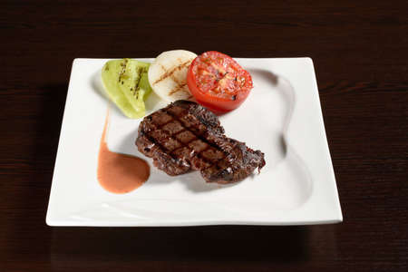 Roasted meat of marble beef steaks with grilled vegetables of tomato, onion and bell pepper on a square plate on a wooden table. Photos for restaurant and cafe menusの写真素材