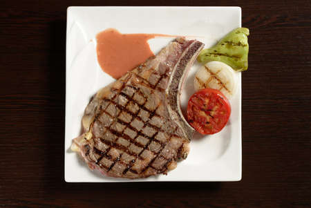 Grilled cowboy beef steak with grilled vegetables of tomato, onion and bell pepper on a square plate on a wooden table. Top view. Photos for restaurant and cafe menusの写真素材