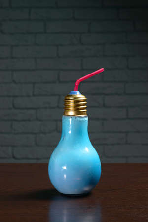 Cocktail in a blue glass in the shape of a light bulb on a wooden table against a brick wall. Photos...