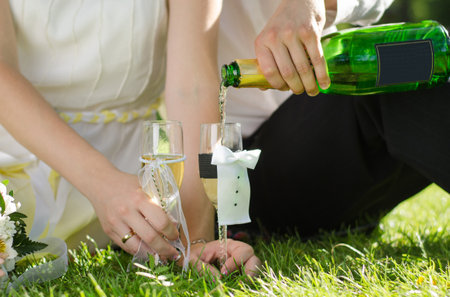 Wedding glasses stand on the grass, the groom's hand pours champagne into the glasses. Close upの写真素材