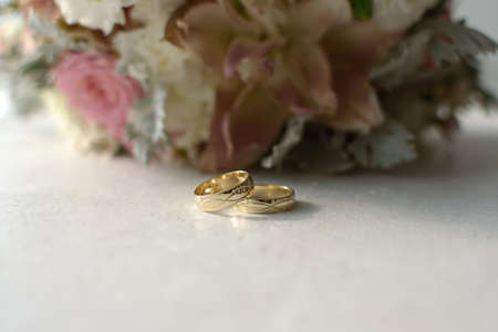 Two gold wedding rings are lying on a white table against the background of a blurry bouquet of flowersの写真素材