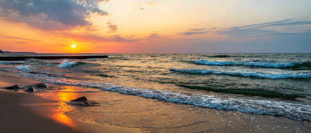 Sunset in the wavy sea with a wooden breakwater and stones on the sandy shore, beautifully colored skyの写真素材