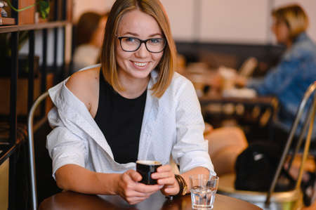 cute girl with a beautiful smile in glasses sitting in a cafe. Drinking coffee, holding a cap of coffeeの写真素材