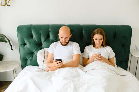 young couple lie in bed with phones. Communication and relationship problems.