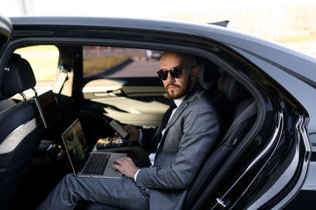Handsome businessman sitting with laptop on the backseat of the car. travelling by a car and working...