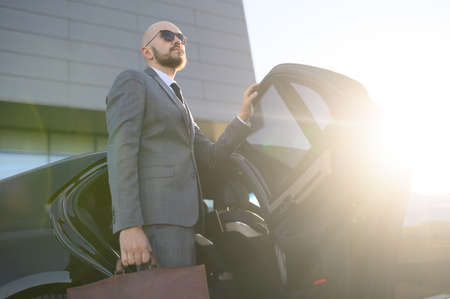 Portrait of a handsome businessman in sunglasses standing near the car outdoors in front of the modern building facadeの写真素材