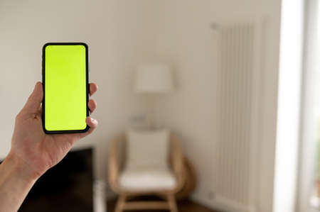 Mockup image of hand holding black mobile phone with blank green screen and background modern interiorの写真素材