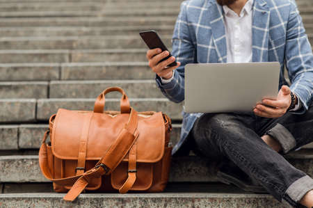 work outdoors, a man sits on the stairs and works on a laptop and talking on the phone. Modern lifestyleの写真素材