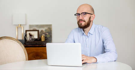 a man in a blue shirt with a beard is working on a laptop at home. Stay in homeの写真素材