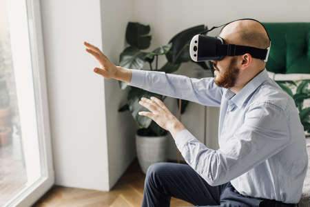 Young Man Wearing Virtual Reality Headset Plays in a Video Game at Home