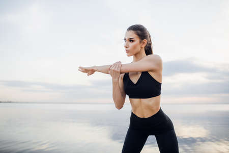 young woman in black sportswear stretching hands by the river. Fitness girl with hands raised above...