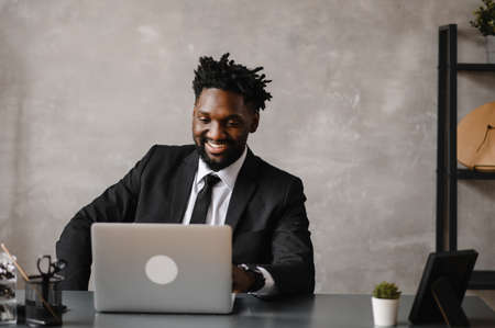 Handsome african american businessman in classic suit uses laptop and smiles while working in office