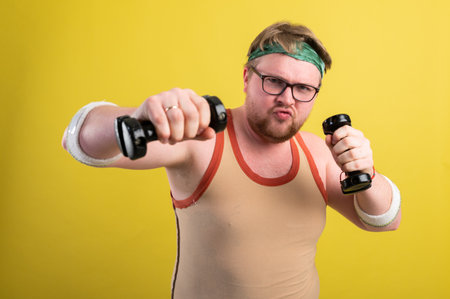 Funny fat man in sportswear with dumbbells in his hands. Overweight man goes in for sports. Yellow background isolateの写真素材