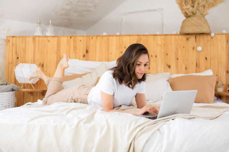 A young businesswoman looks at the screen of a laptop working on the Internet. The girl is lying on the bed and using a laptopの写真素材