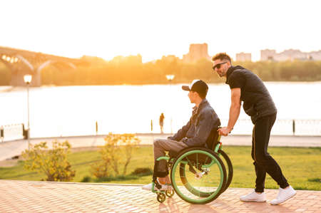 a man in a wheelchair walks with his friend. Man walking with disabled friend in wheelchair at parkの写真素材