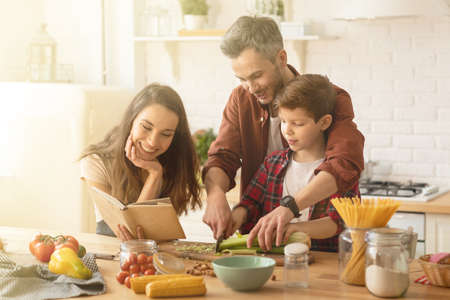 Caring parents and child cooking. father teaching little son use knife. Toothy smiling mother holding cookbook with salad recipe. Home kitchen interior with fresh healthy vegetables food on tableの写真素材