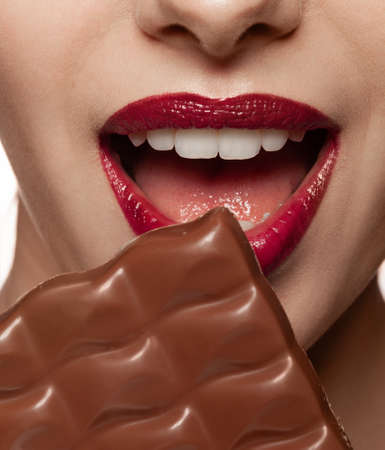 Female open mouth and chocolate bar closeup shot. Beautiful women lips with red burgundy lipstick makeup and white teeth. Girl tasting, eating, biting sweets. Detail face and brown milky dessertの写真素材