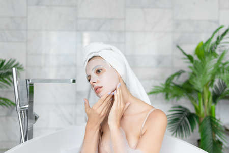 Woman wearing towel on head applying face sheet mask while taking bath with soap foam.の写真素材