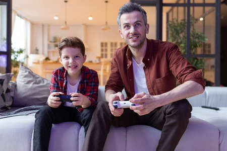 Father and son playing video game in home living room. Dad and child sitting on sofa hold console...