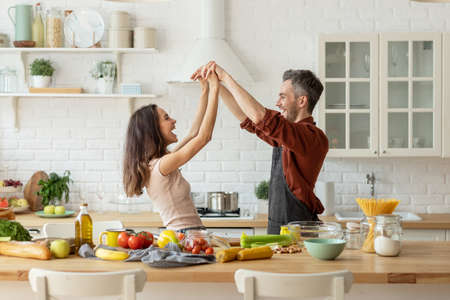 Happy laughing wife and husband dancing while cooking in bright loft on home kitchen. Young bearded man holding woman hands standing behind table with fresh food ingredients. Excited loving coupleの写真素材
