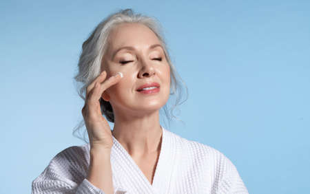 Senior woman with eyes closed putting cosmetic moisturizing rejuvenating cream on face feeling pleasure. Studio portrait on blue. Refreshment and anti-wrinkle skin therapyの写真素材