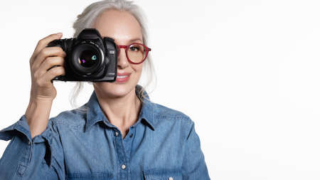 Smiling stylish senior woman with trendy outfit take photo on digital camera portrait on white. Elderly photographer amateur or professional. Tourism and hobby. Best vibes, good memoryの写真素材