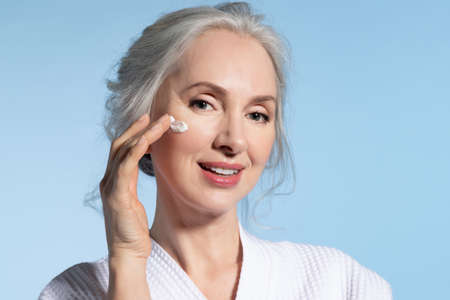 Beautiful senior woman wearing bathrobe applying daily skin care moisturizing nourishing facial...