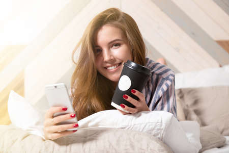 Smiling teen girl wearing pajamas with phone and coffee lying in bed looking at camera closeup...
