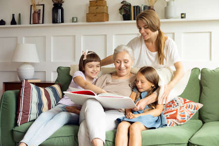 Mom covers grandmother and granddaughters reading a book with a cozy blanketの写真素材