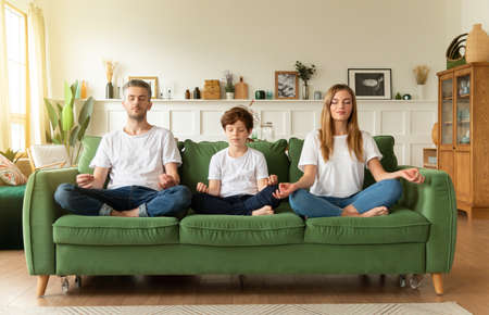 Calm young family with son practice yoga together sitting in Lotus position on couch. Conscious parents with school boy child rest on sofa meditating relieve negative emotions on weekend at home.の写真素材