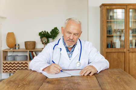 Serious mature doctor in white uniform with stethoscope on neck makes notes in medical journal sitting at desk. Busy senior therapist practitioner gp writing illness history, filling patient card.の写真素材