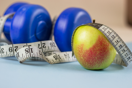 Blue dumbbells, measuring tape and apple on a blue background. Healthy lifestyle, the concept of body weight loss. Cares about the body. Empty place for text.の写真素材