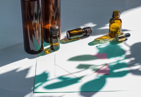 Dark cosmetic bottles and green natural leaves on a light background. Green empty card, sheet for writing. Layout for adding inscriptions. Natural hard light, deep shadows. The concept of natural environmentally friendly cosmeticsの写真素材