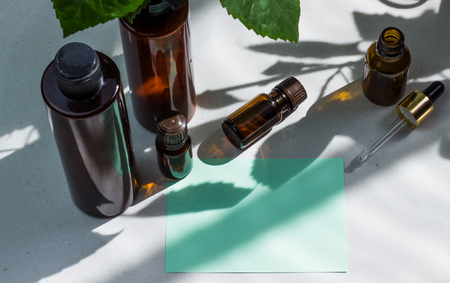 Dark cosmetic bottles and green natural leaves on a light background. Green empty card, sheet for writing. Layout for adding inscriptions. Natural hard light, deep shadows. The concept of natural environmentally friendly cosmeticsの写真素材
