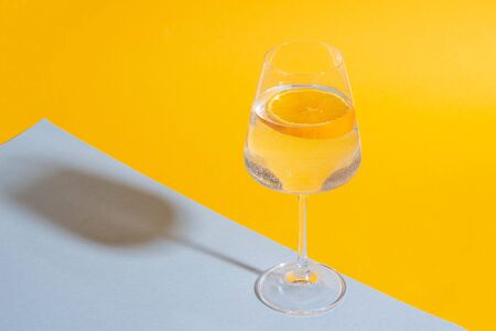 Glass with a summer drink and a slice of orange on a bright yellow and blue background with bright light and harsh shadowsの写真素材