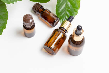 Glass cosmetic bottles with a dropper stand next to green leaves on a white background. Organic cosmetics concept, natural essential oil and creamの写真素材