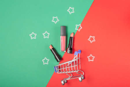 Women's lipsticks and lip glosses in a shopping trolley on a bright red and green background. The concept of buying cosmetics, online shopping, holidayの写真素材