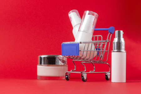 A set of face and body cosmetics and a shopping trolley on a bright red background. The concept of buying cosmetics, online store, holidayの写真素材